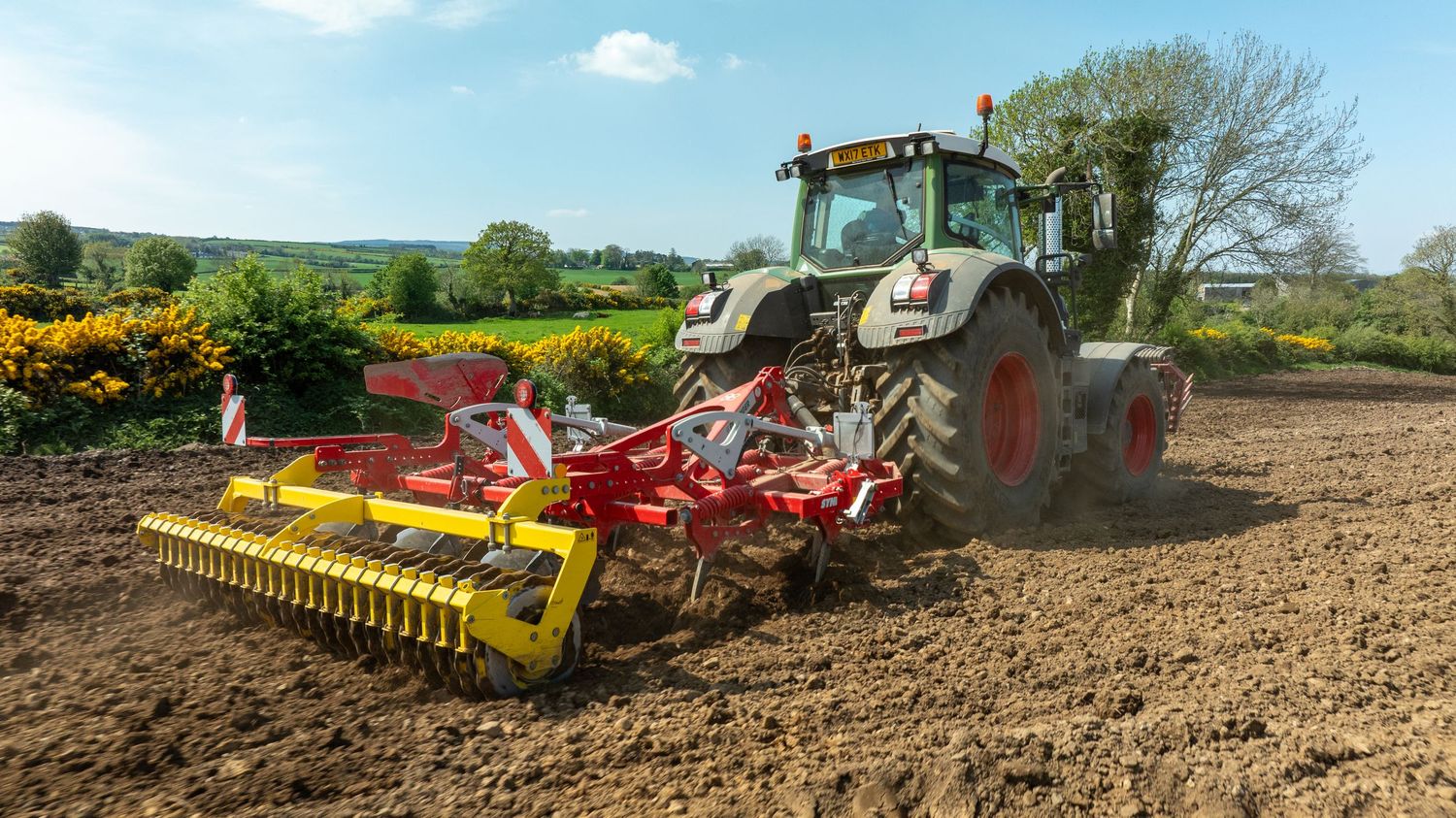 Modern tractor pulling soil cultivation equipment across a freshly plowed field on a sunny day in rural countryside.