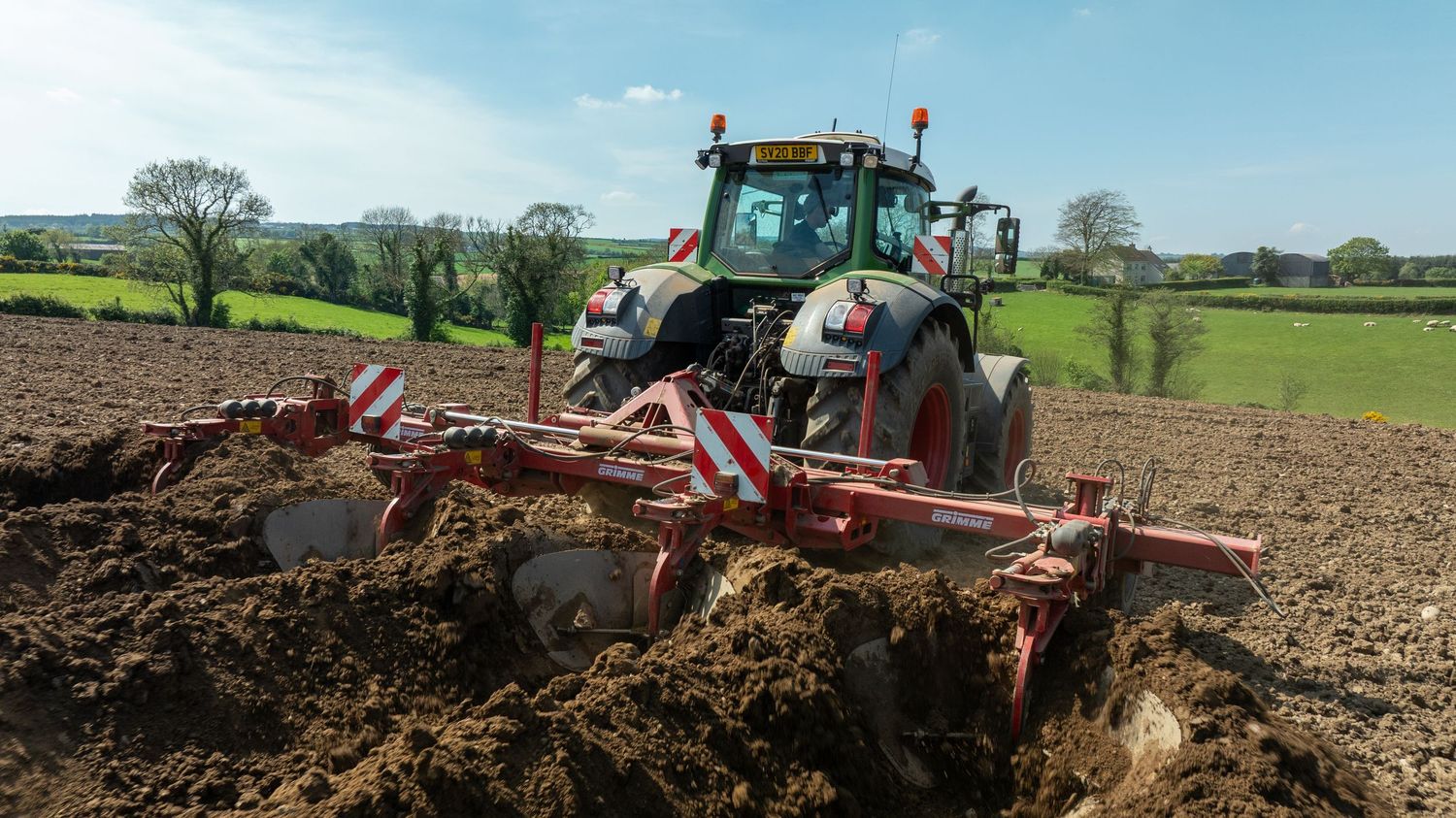 A green tractor pulling a large cultivator implement through brown soil in a rural agricultural field.