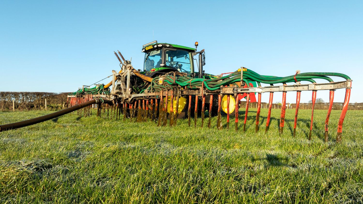 Large agricultural machinery with red and green implements stretches across a grass field against a blue sky.