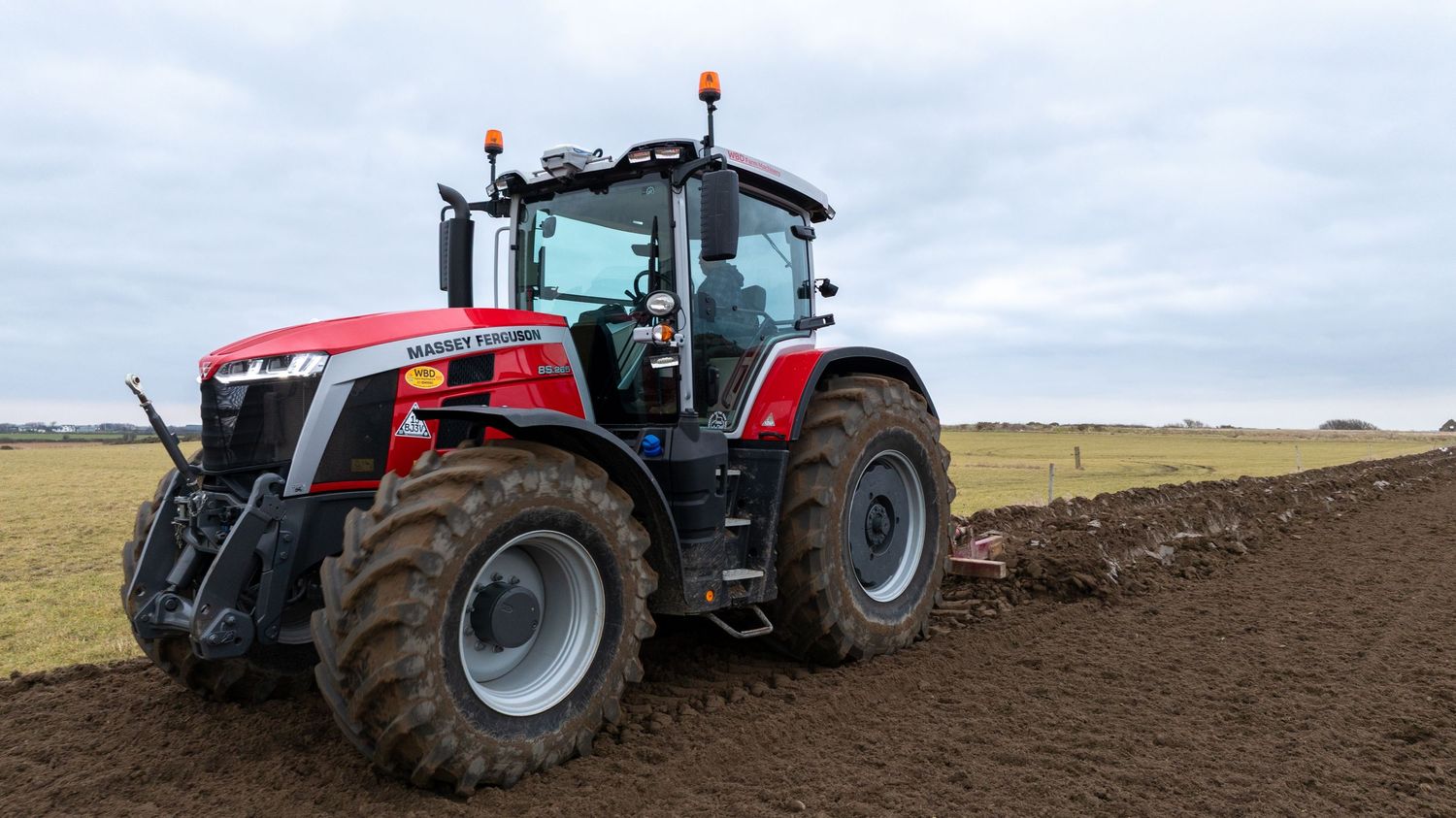 Modern red Massey Ferguson agricultural tractor plowing field on farmland under cloudy sky.