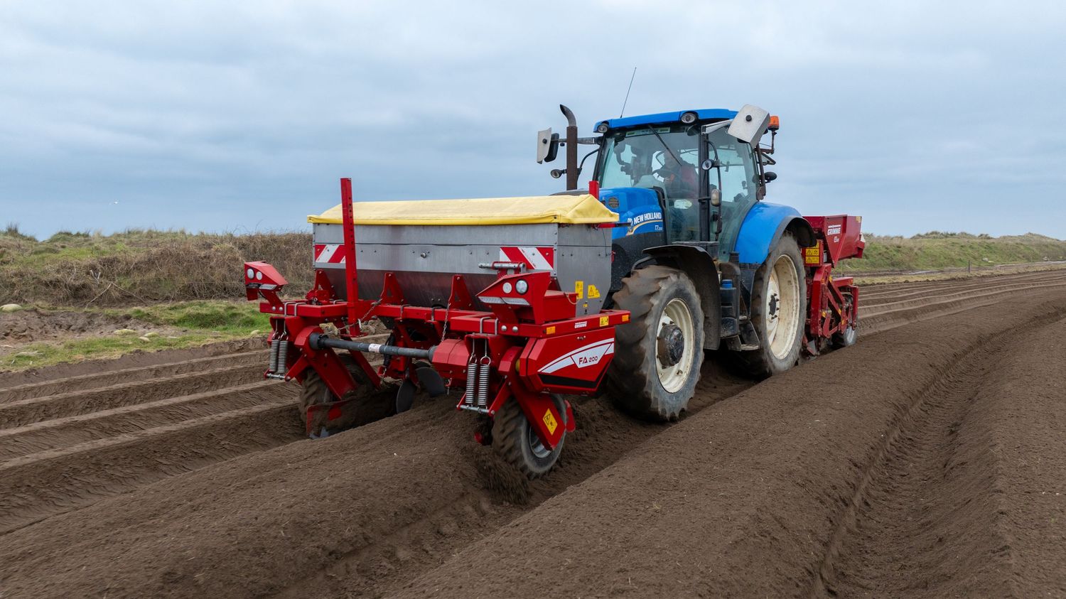 Blue tractor with red planting equipment working in a freshly tilled agricultural field under cloudy skies.