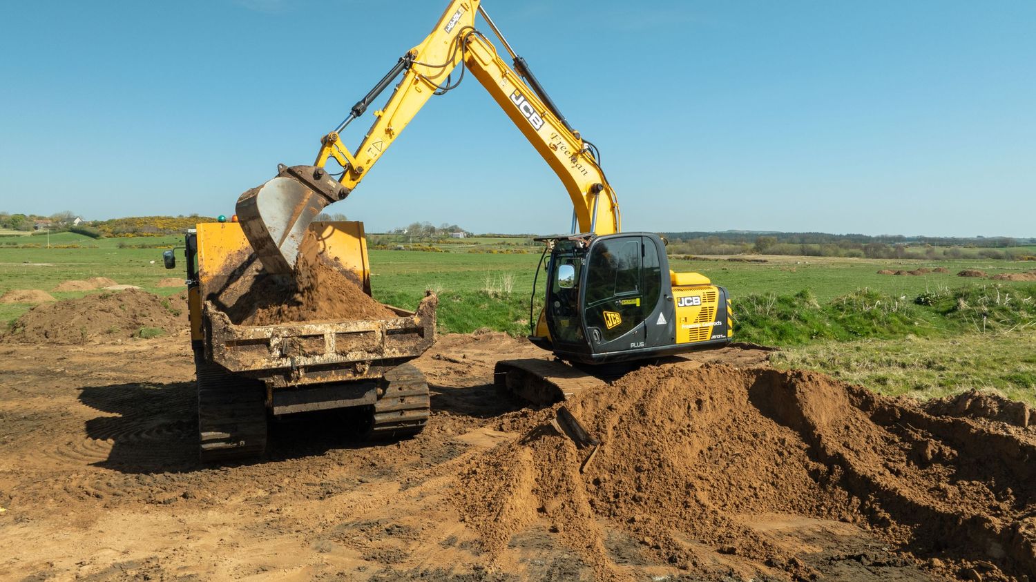 Yellow excavator loading soil into a dump truck at a construction site with green fields in the background.