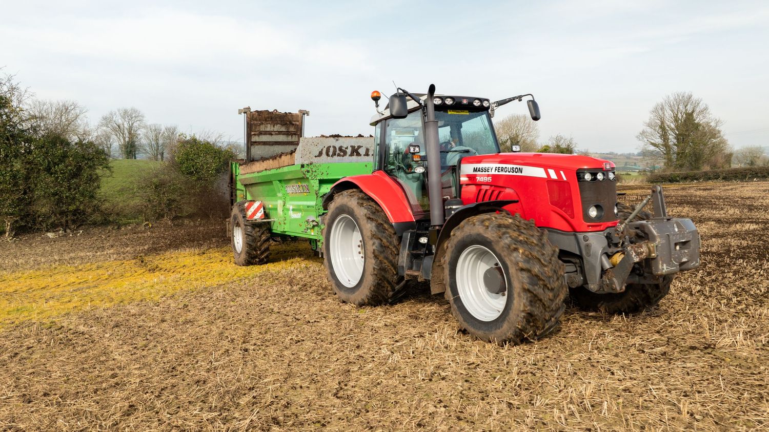 A red Massey Ferguson tractor pulling a lime green fertilizer spreader across a brown field on a cloudy day.