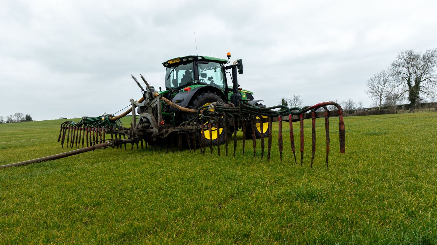 Green tractor with tillage equipment operates in a grassy field under cloudy skies.