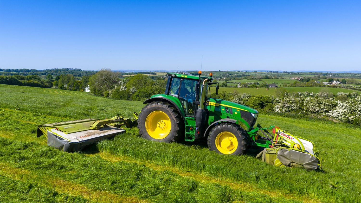 John Deere tractor with mowing attachments working on rolling green pastureland against a blue sky background.