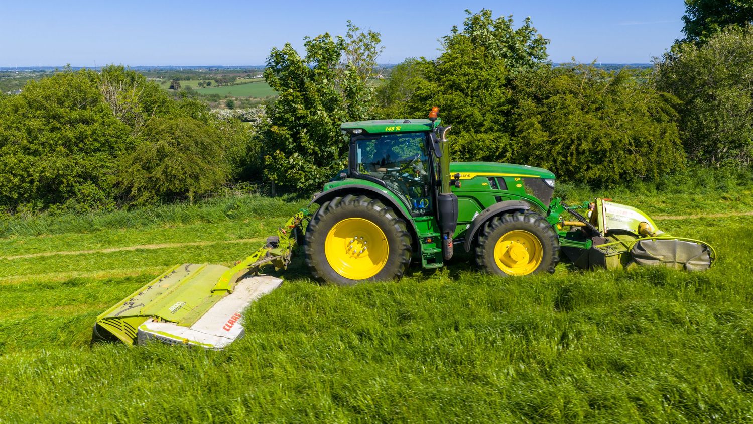 Green John Deere tractor mowing tall grass on a hillside with mower attachments on a sunny summer day.
