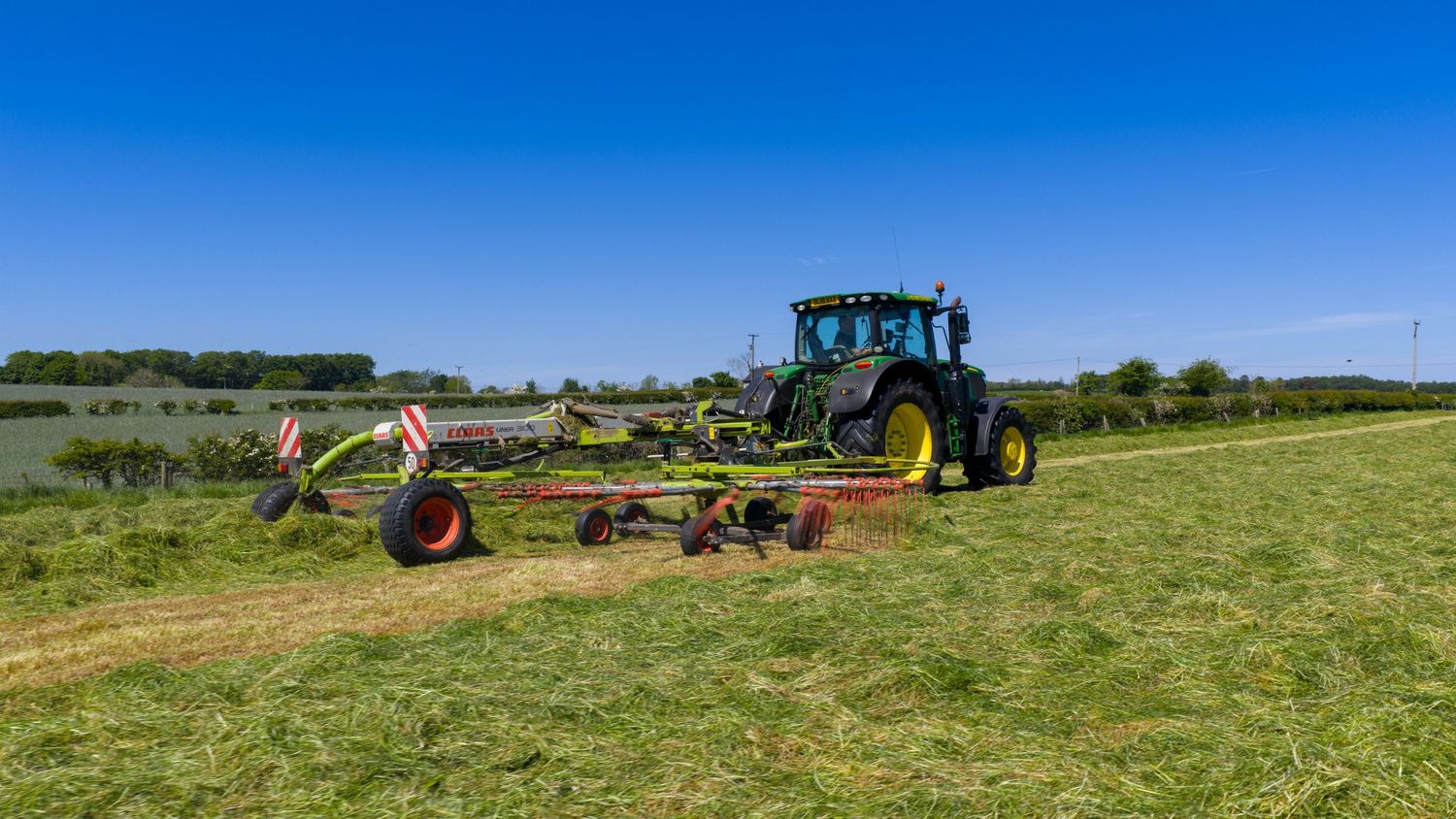 John Deere tractor with multiple hay raking implements working in a grassy field under blue skies.