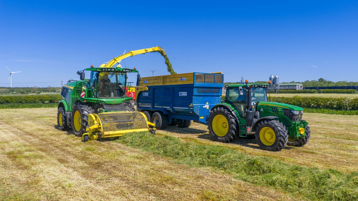 Two green John Deere tractors working in a field with one harvesting hay and transferring it to a blue trailer.