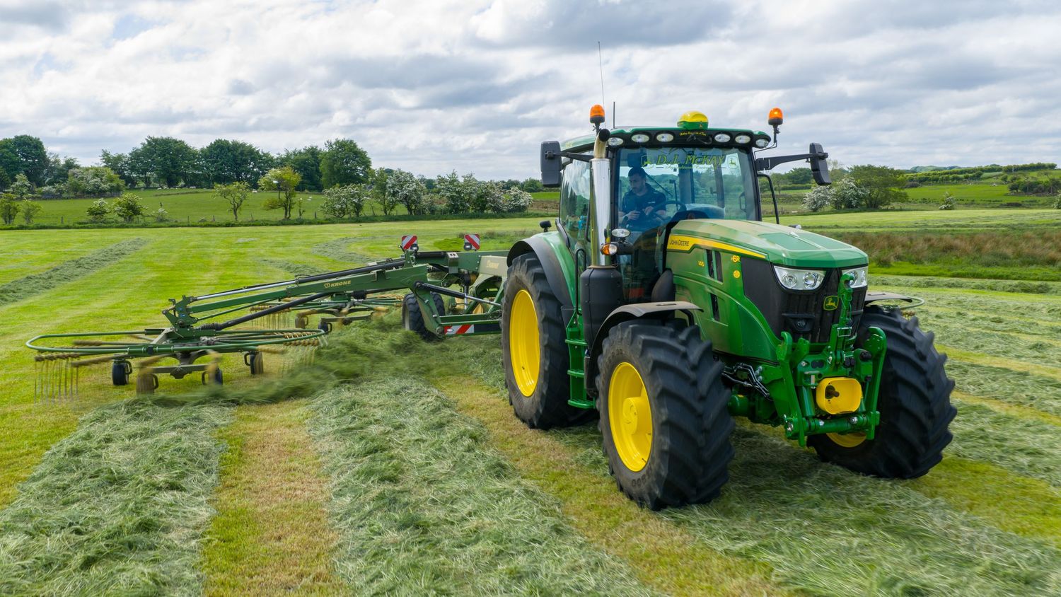 John Deere tractor with green hay turner attachment working on a lush grass field under cloudy skies.