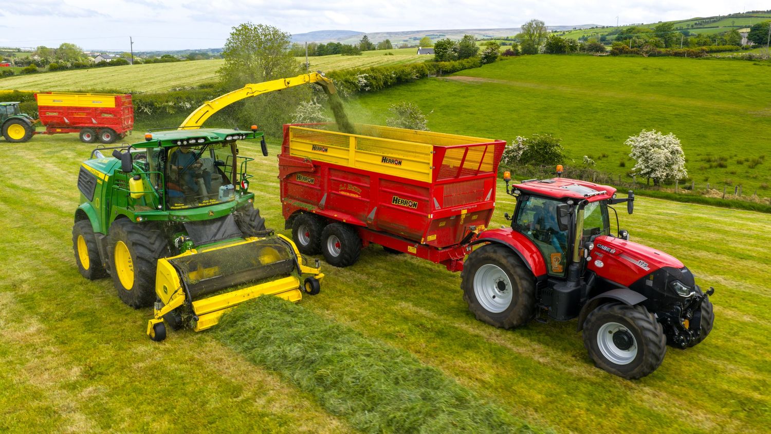 Green forage harvester collecting and loading fresh cut grass into a red trailer being pulled by a red tractor in rolling fields.