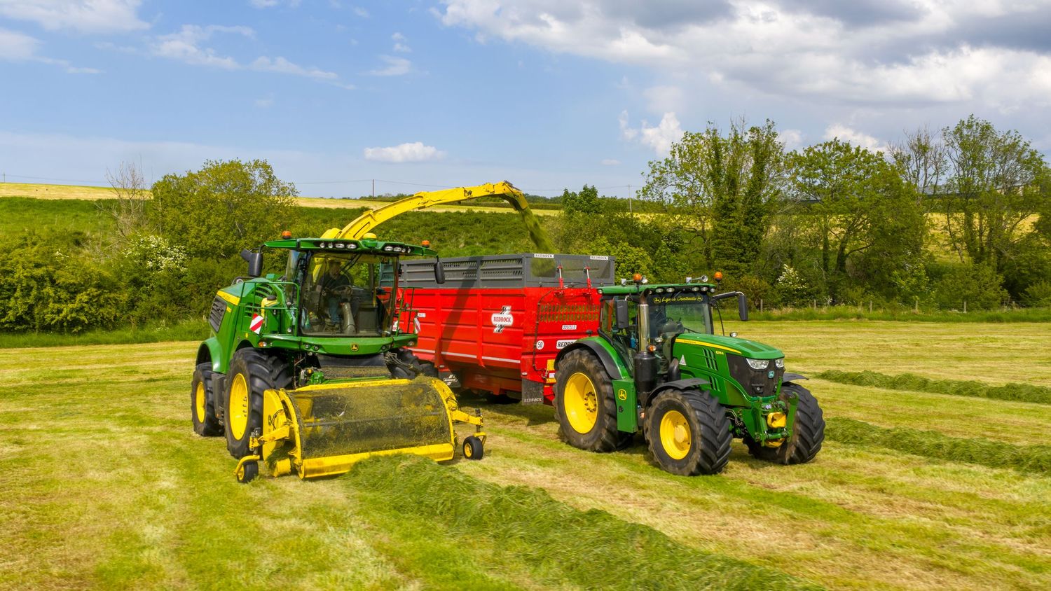 Modern John Deere tractors and farm equipment collecting hay from a green field under blue skies with white clouds.