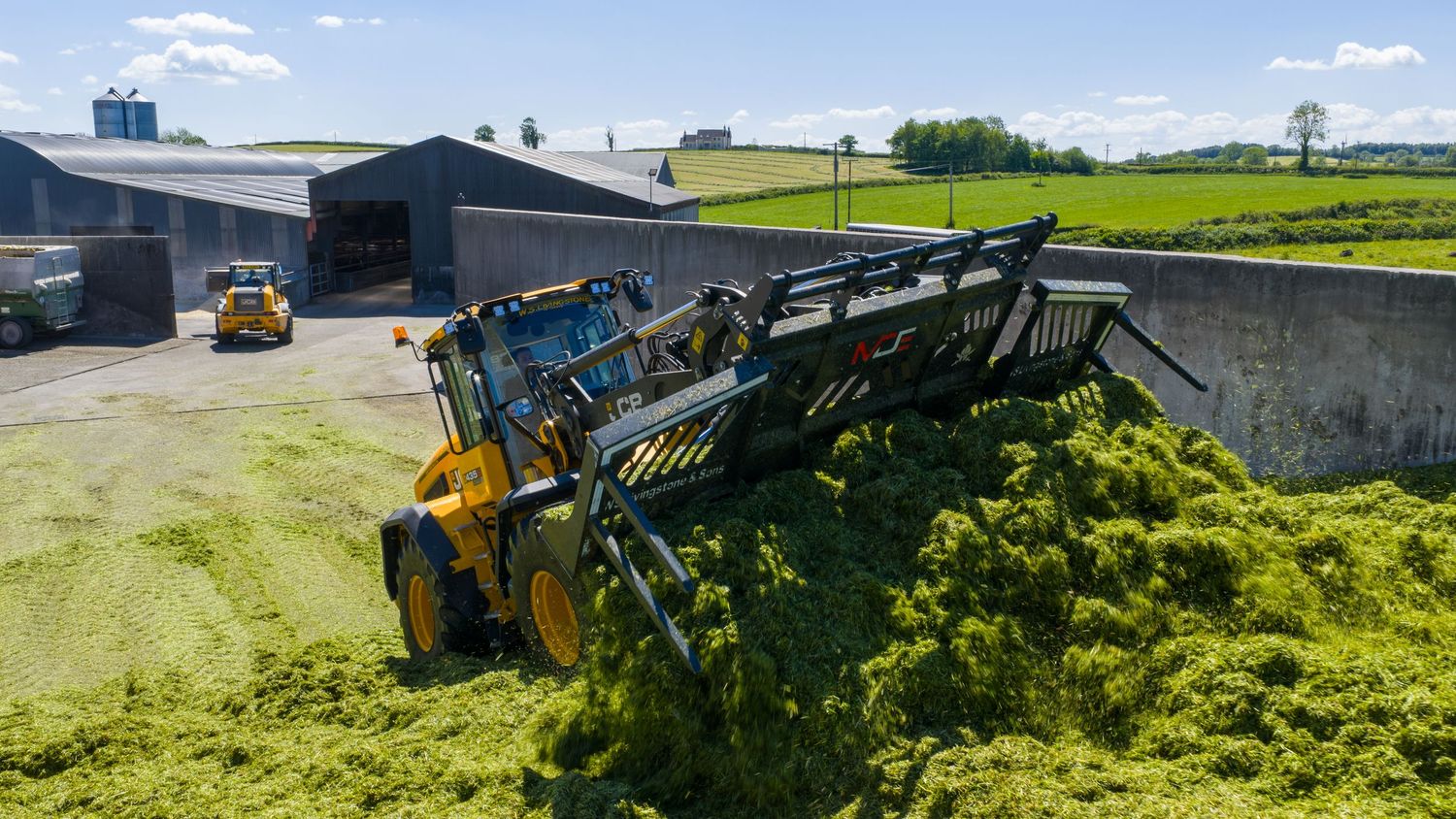 Yellow front loader tractor moving silage at a dairy farm with green fields in the background.