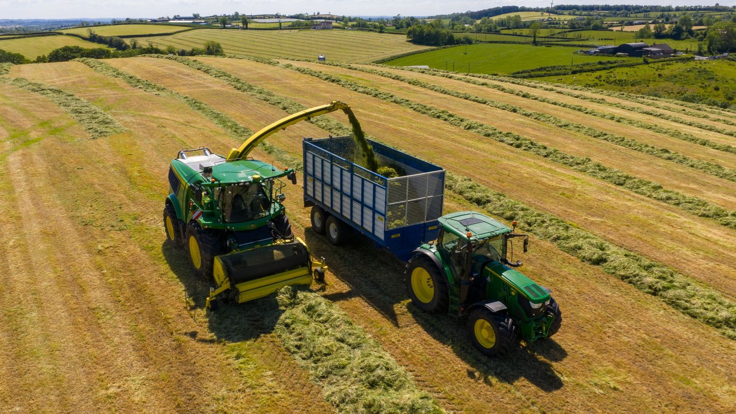 Aerial view of farming equipment harvesting hay with a forage harvester loading into a trailer pulled by green tractor.