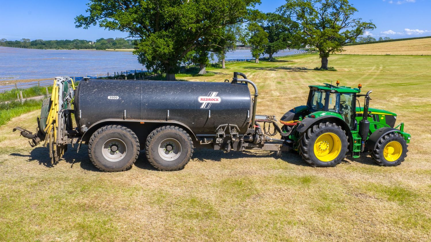 John Deere tractor pulls black liquid manure fertilizer tank across green lakeside field on sunny day.