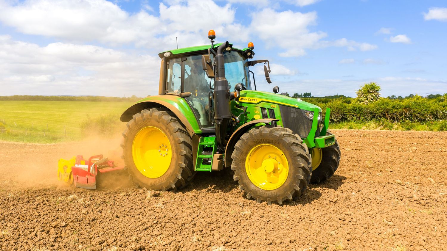 Modern green John Deere tractor plowing dry agricultural field on sunny day with blue sky and clouds.