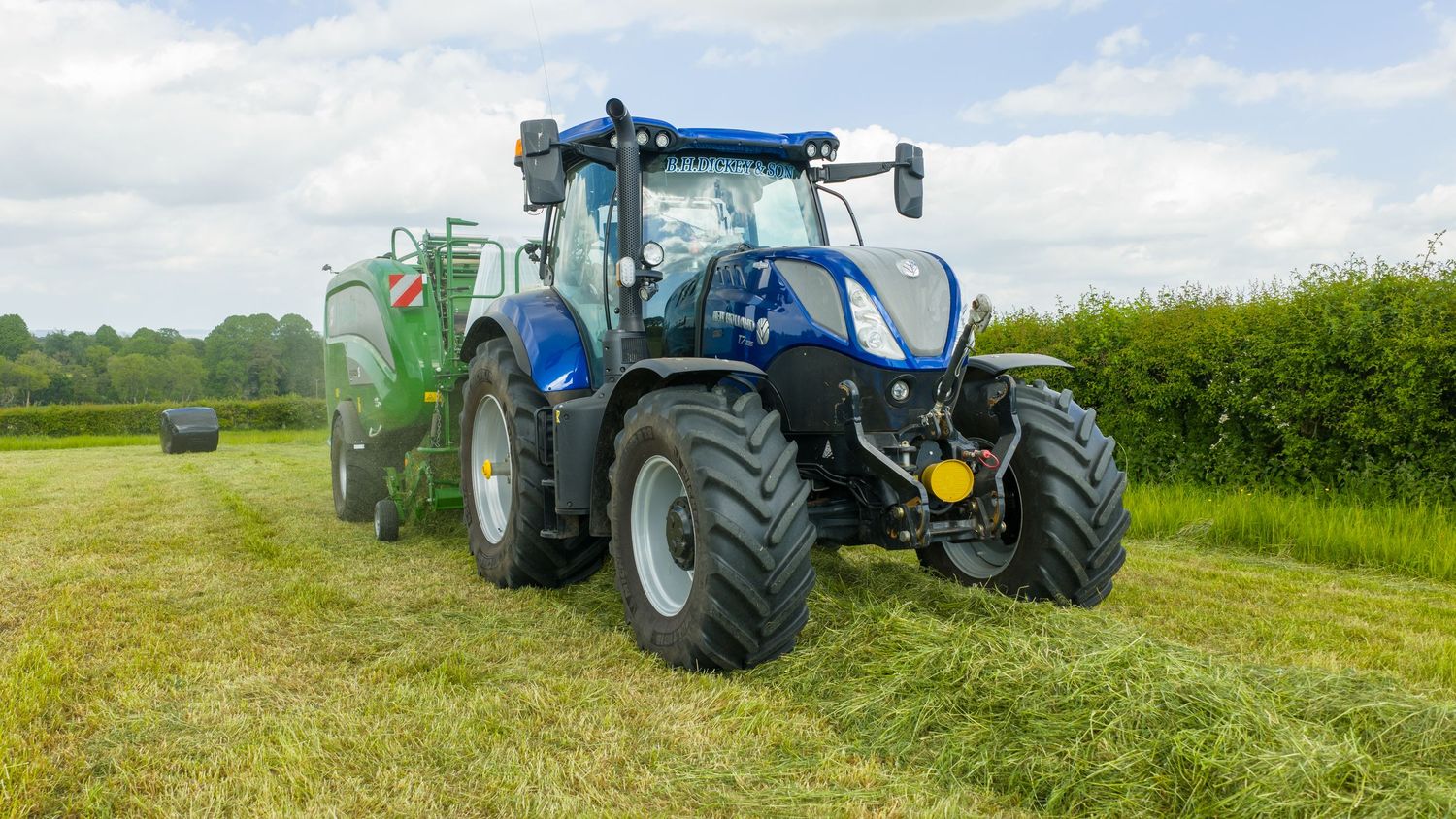 Blue New Holland tractor with liquid storage tanks attached working in green agricultural field on cloudy day.