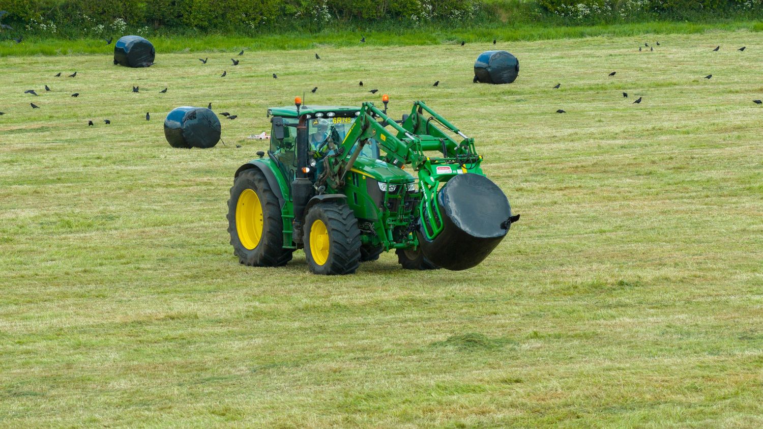Green John Deere tractor carrying hay bales on mowed farmland field with round bales scattered in background.