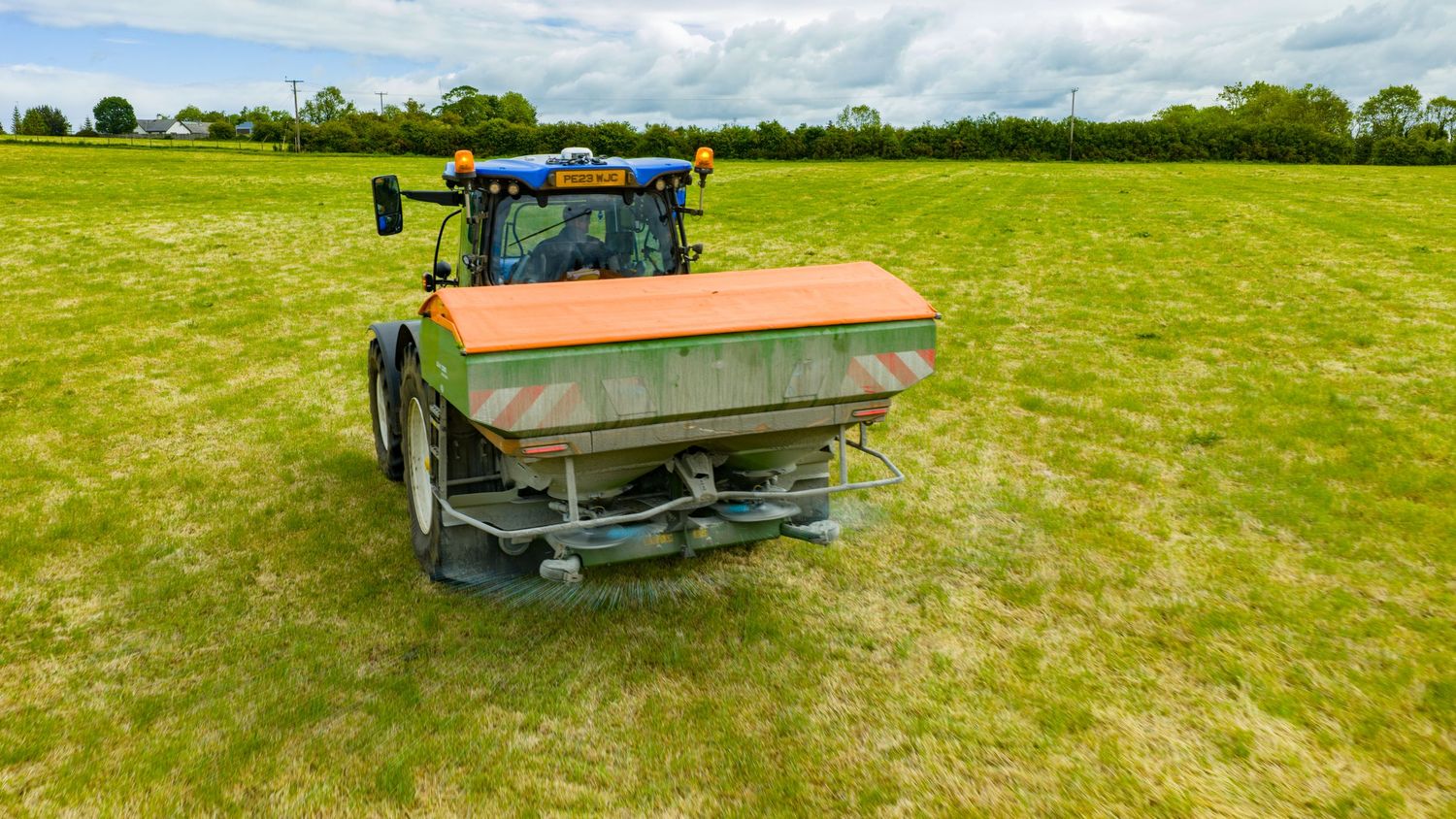 A green tractor with fertilizer spreader equipment operates in a lush agricultural field under partly cloudy skies.