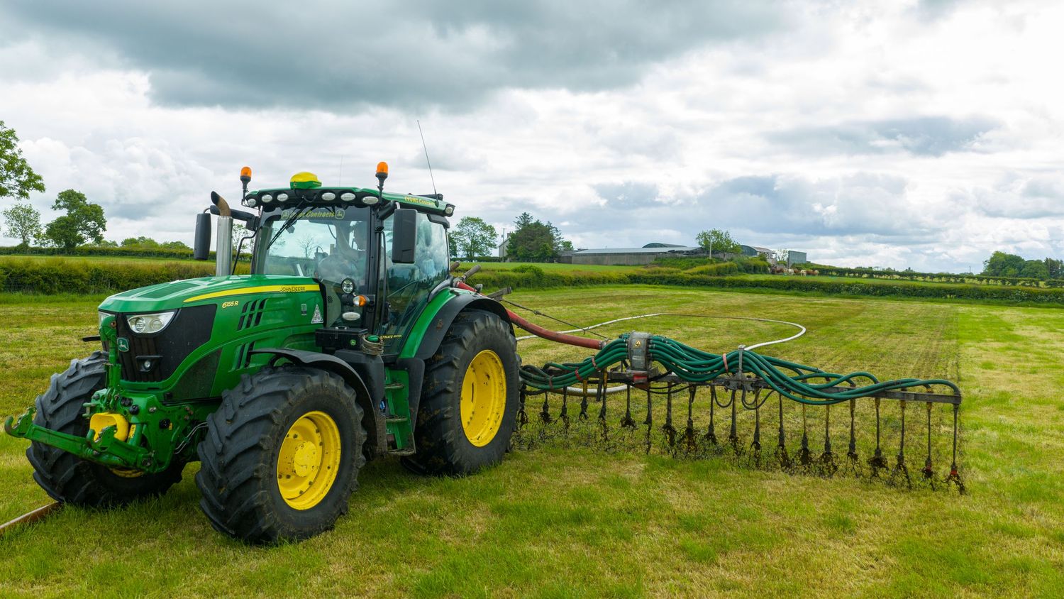 A green John Deere tractor with attached hay rake equipment working in a lush grass field under cloudy skies.
