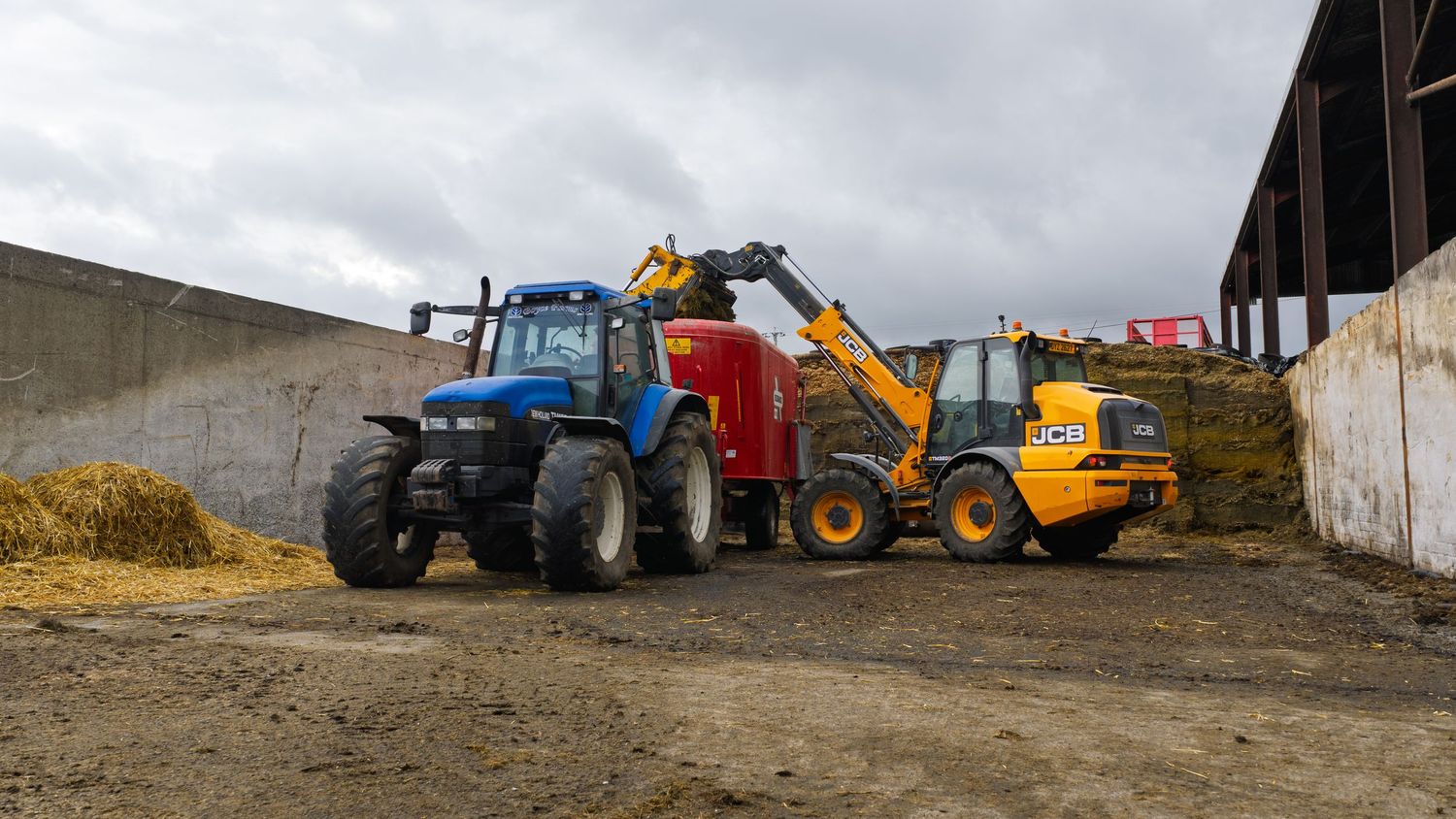 Agricultural equipment including blue tractor and yellow loader working together near silage storage area under cloudy sky.