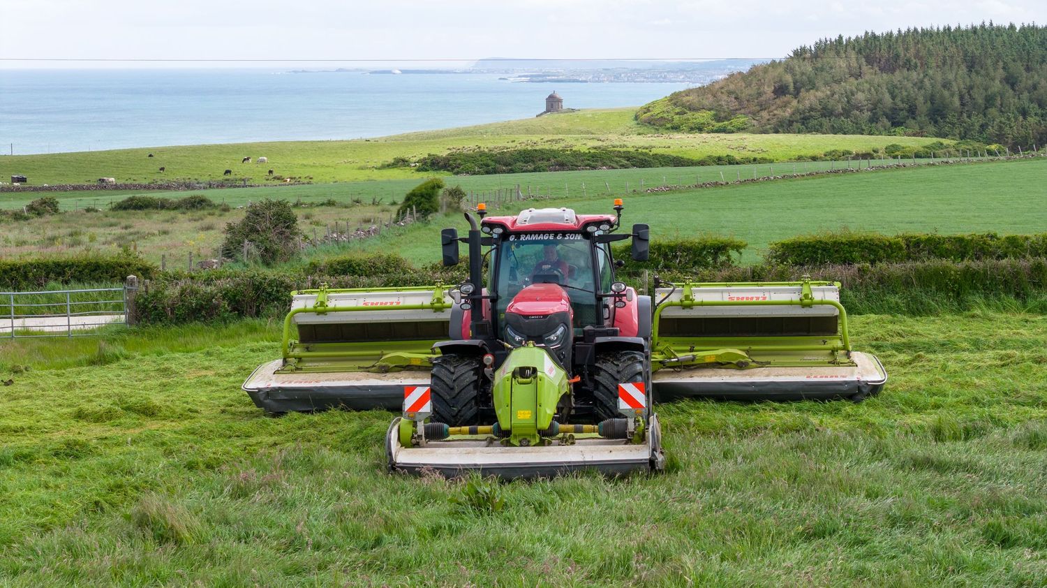 Red tractor with twin mowers cutting grass on a lush green hillside overlooking the ocean.