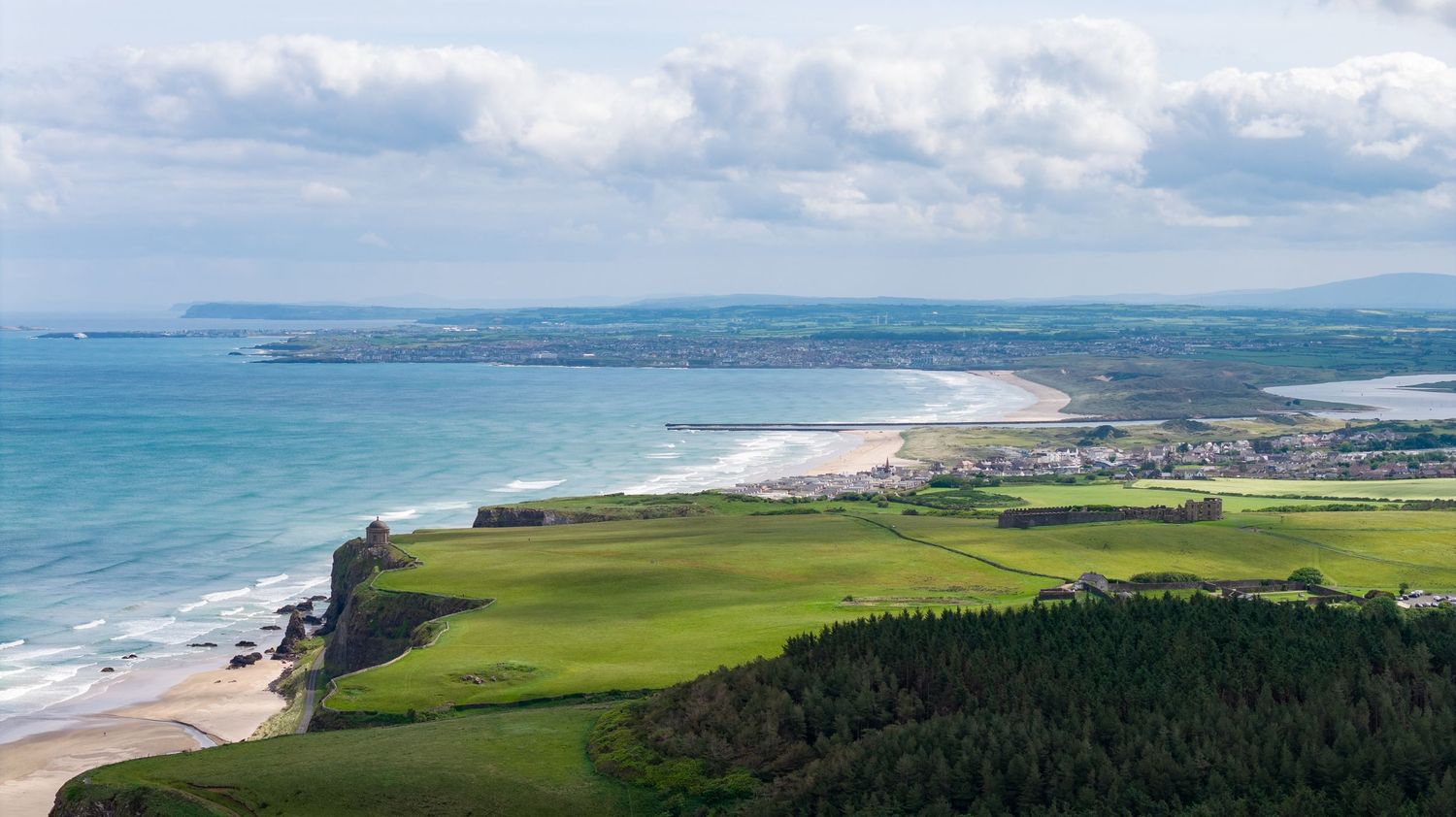 Aerial view of dramatic coastal cliffs with green fields overlooking turquoise waters and sandy beaches in Ireland.