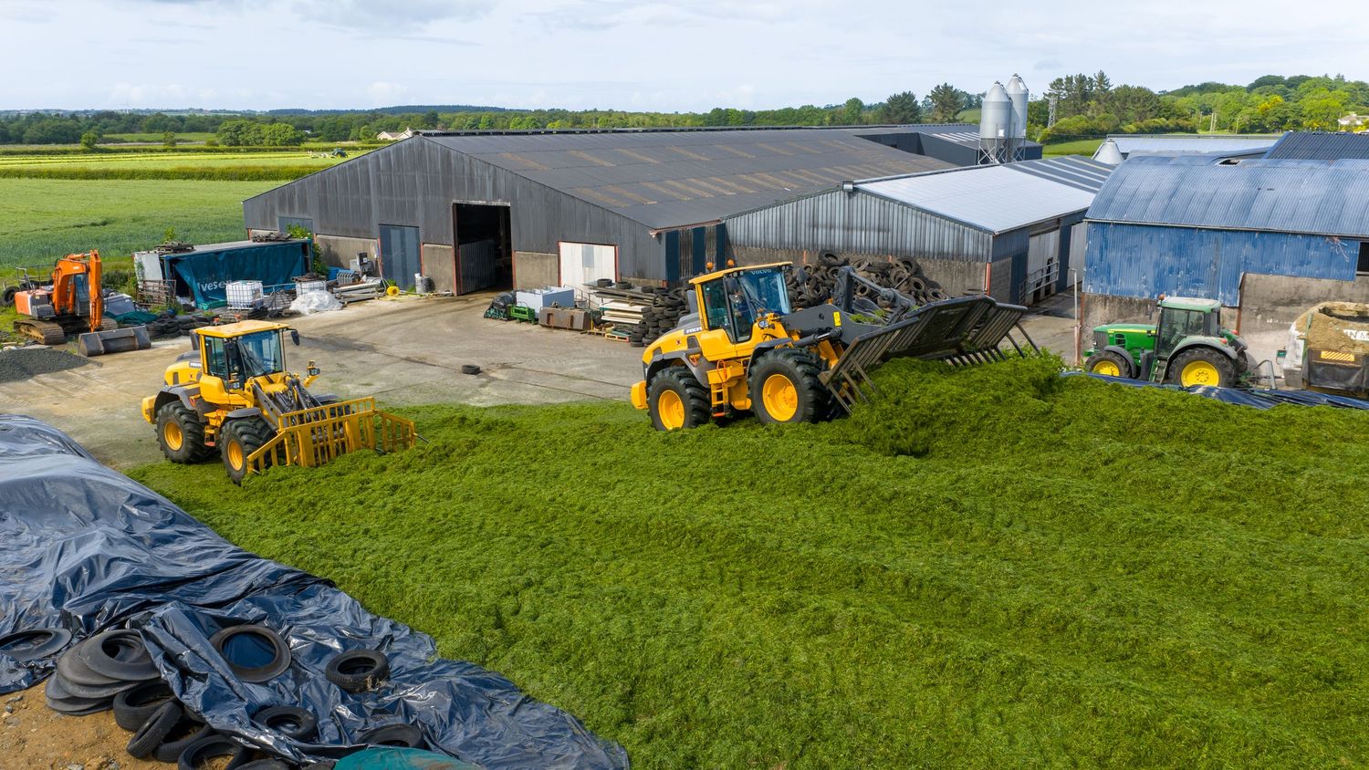 Yellow tractors and machinery work to compact silage at a farm storage facility with grey buildings and green fields.