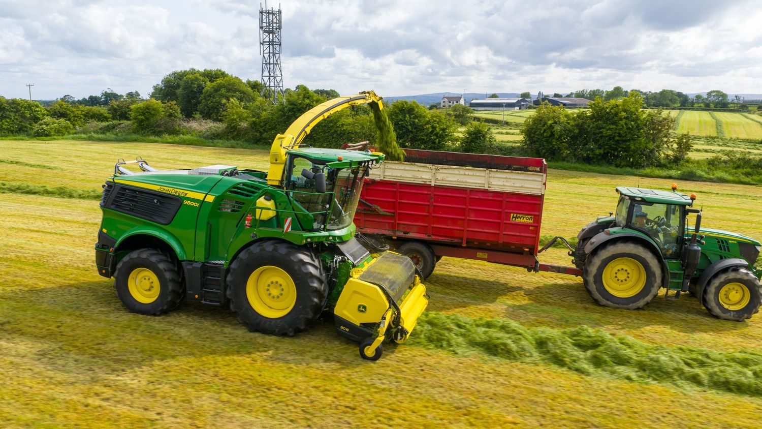 Green farm tractors and equipment working together in a grassy field during harvest season with overcast sky.