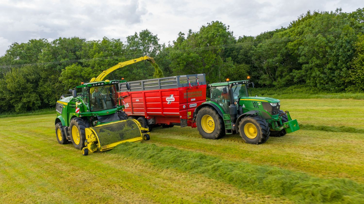 Agricultural machinery harvests grass silage with John Deere forage harvester loading into red trailer pulled by tractor.