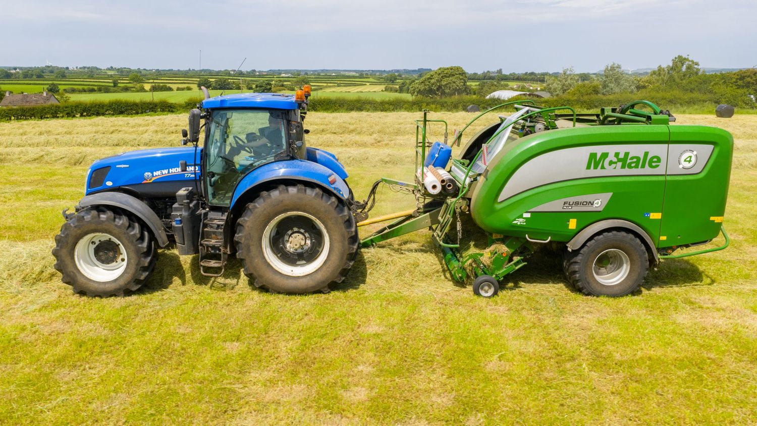 Blue New Holland tractor pulling a green McHale baler in a sunny hay field with rolling hills in the background.