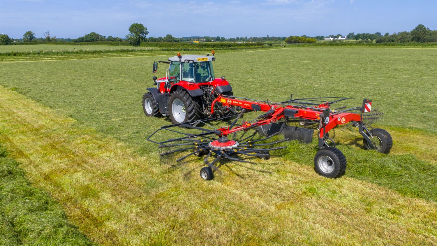 Red tractor pulling a hay rake implement across a green field to gather dry grass for baling.