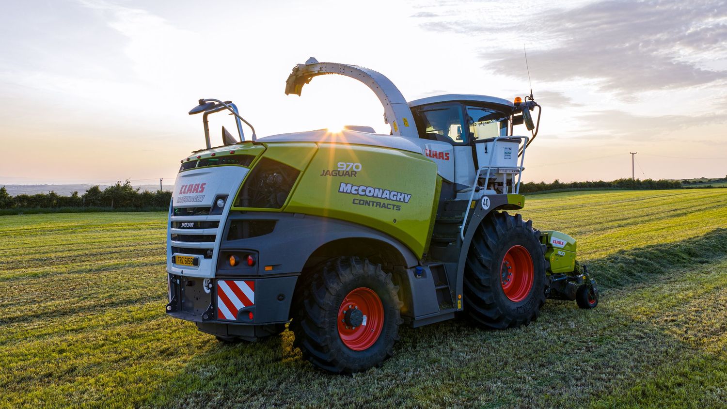 A Claas Jaguar forage harvester operates in a green agricultural field at sunset with vibrant yellow and blue colors.