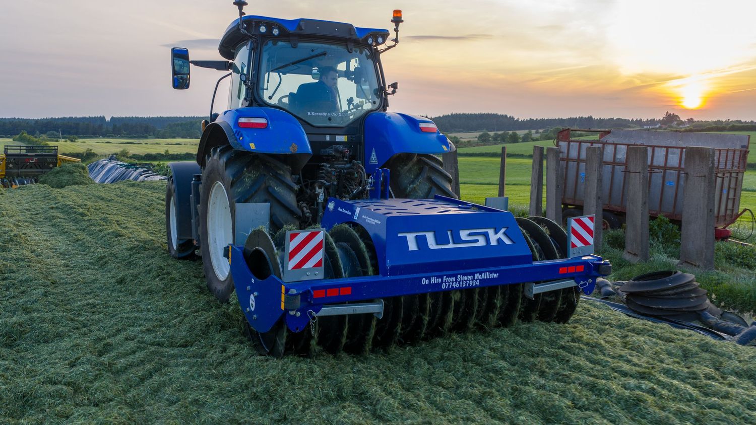 Blue agricultural tractor pulling farm equipment across green fields during a vibrant sunset over rolling countryside.