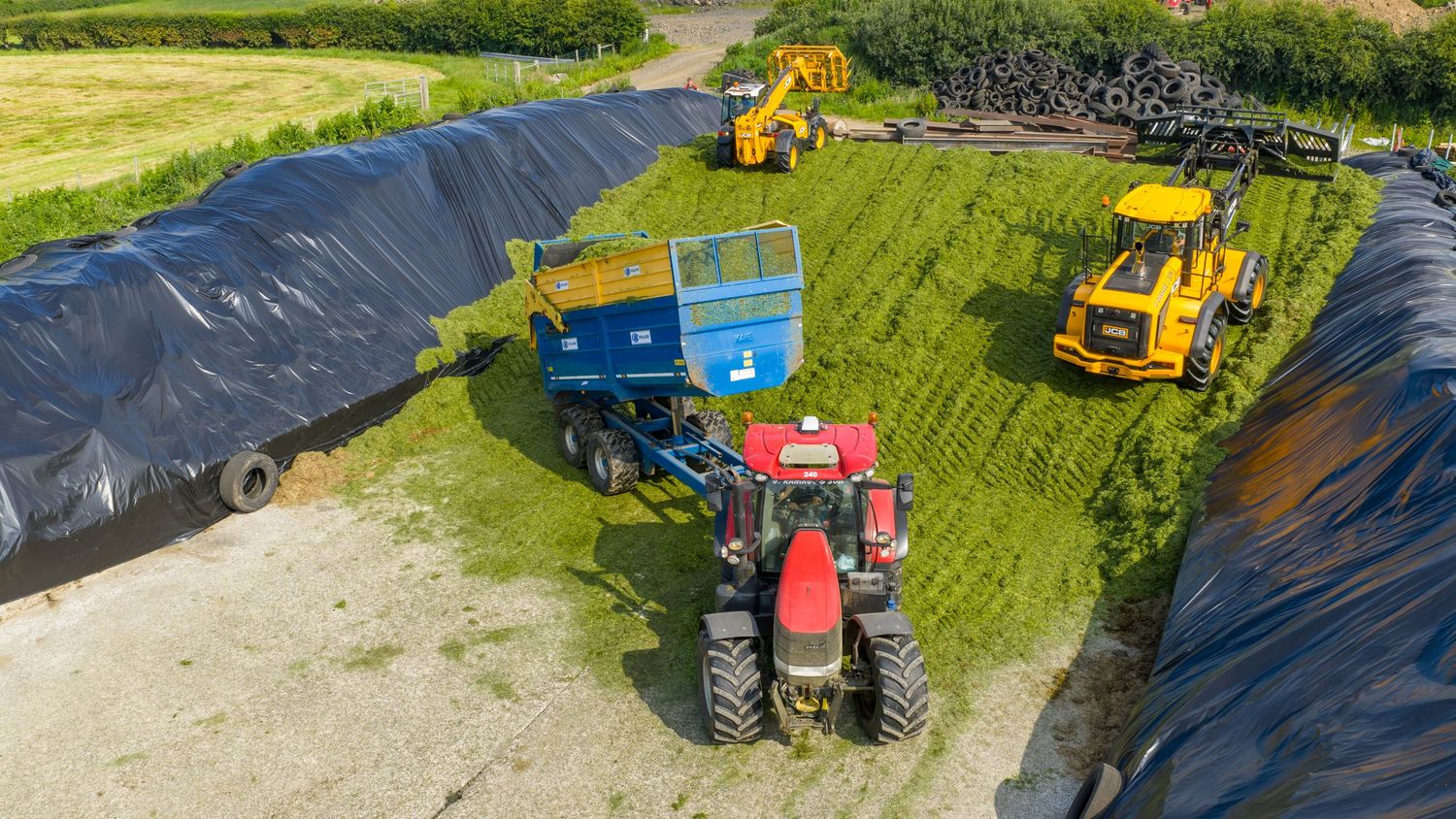 Several agricultural machines and tractors drive in a row across lush green fields during a farming operation in daylight.