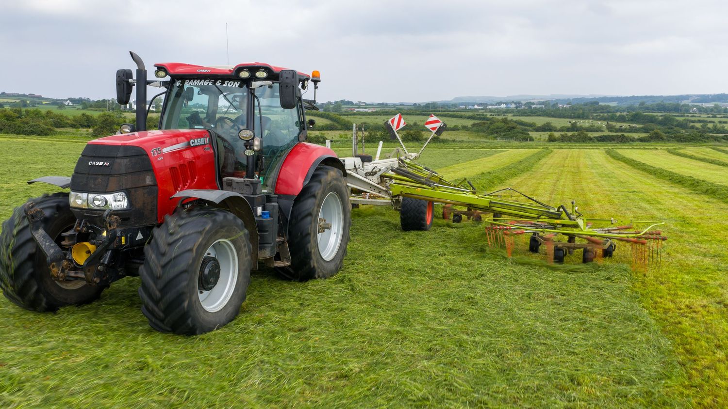 Red tractor pulling modern farming equipment across lush green agricultural fields on a sunny summer day.