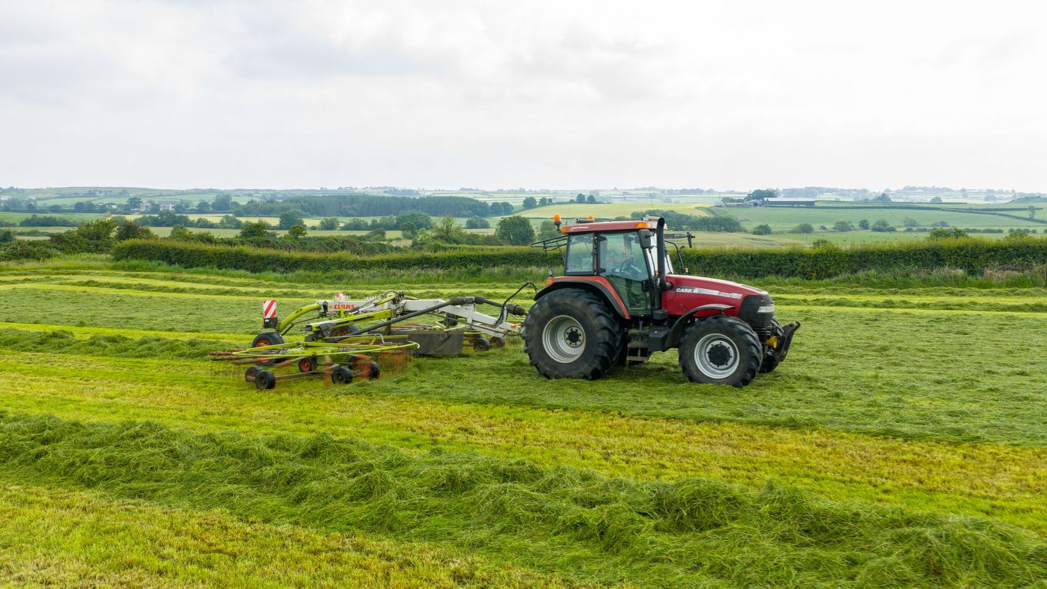 Red tractor mowing green farmland with rolling hills and countryside landscape in the background.