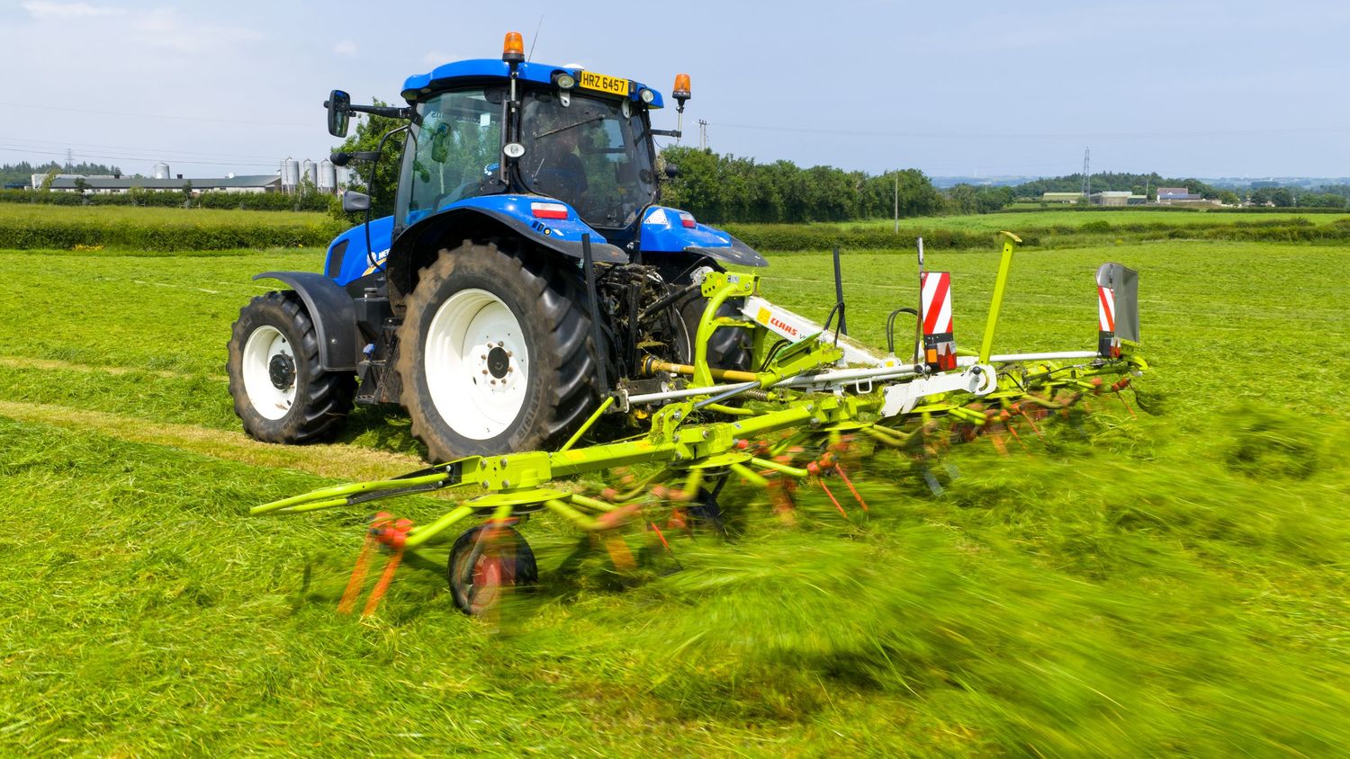 Blue New Holland tractor pulling hay equipment across a vibrant green field during harvest season.