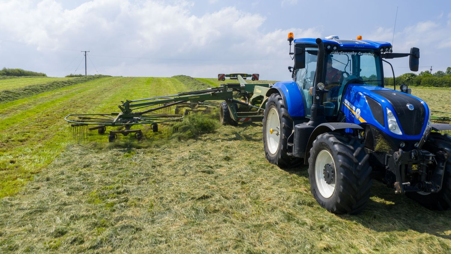 Blue New Holland tractor with hay rake attachment working on green hillside field under blue sky.