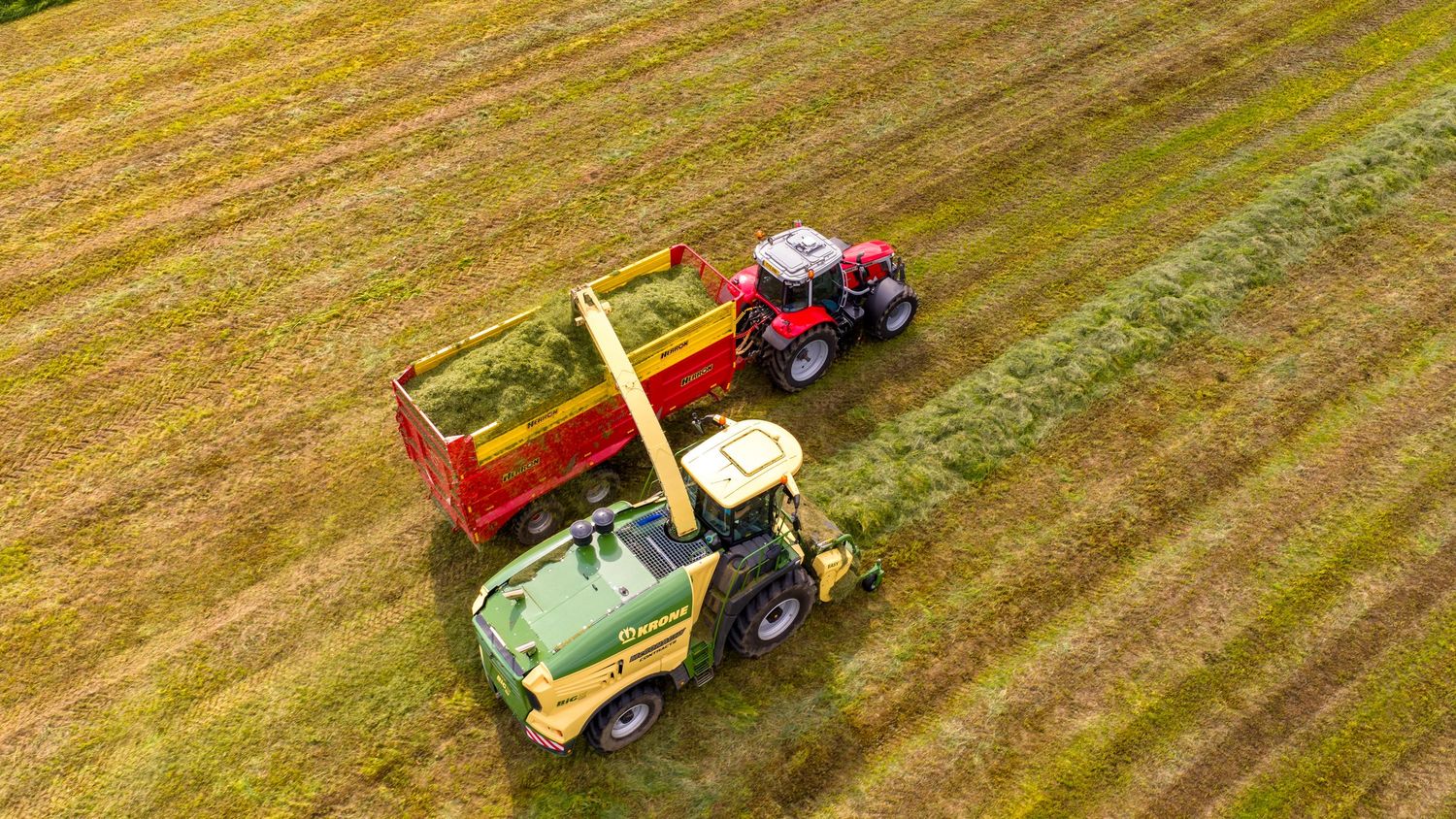 Aerial view of two tractors working in a harvested hay field with one pulling a red wagon trailer.