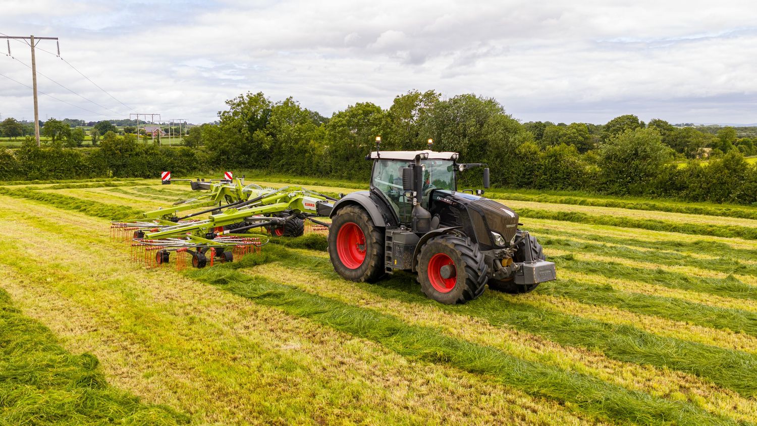 Various tractors with machinery harvest hay in green agricultural fields under cloudy skies during summer season.