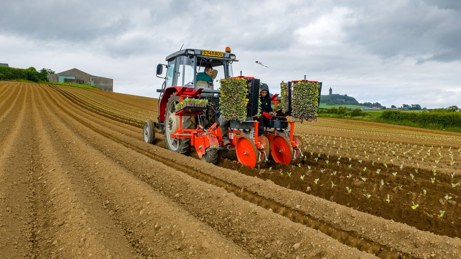 A panoramic view of farm machinery plowing brown agricultural fields under cloudy skies on rolling countryside.
