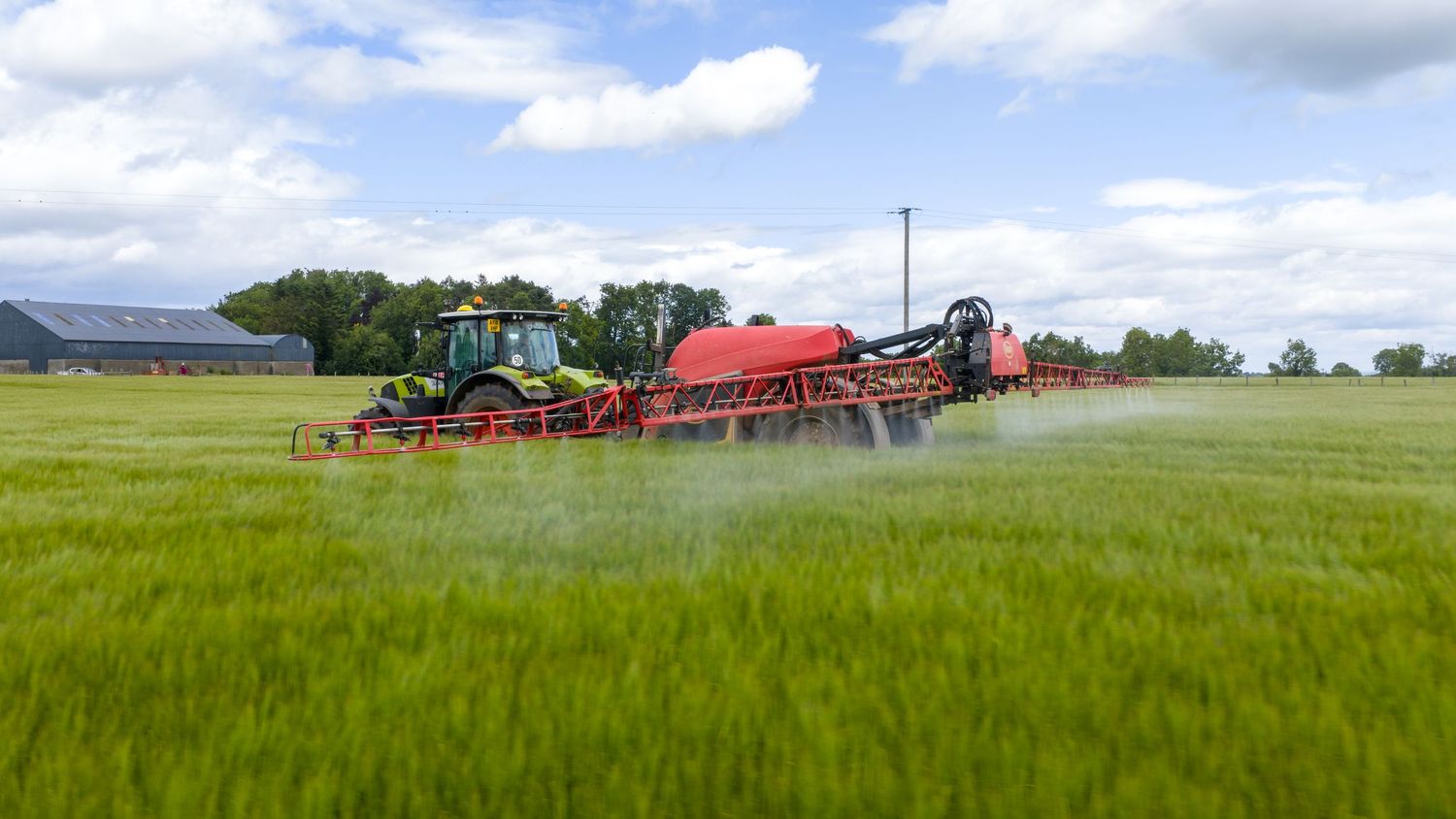 Agricultural tractor pulling sprayer equipment through green wheat field under cloudy sky.