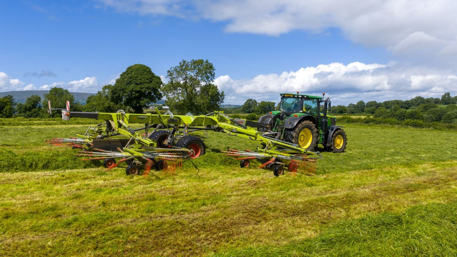 Green John Deere tractor pulling hay raking equipment across a lush grass field on a sunny summer day.