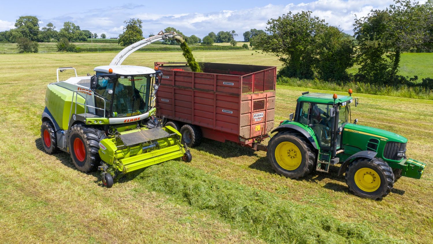Green forage harvester and John Deere tractor with red trailer working together in a lush grass field on sunny day.