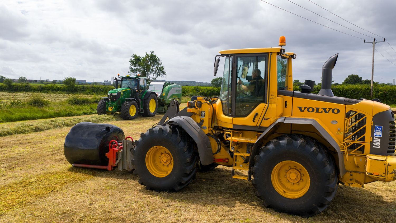 Road grader machinery and tractors parked along a dirt path in a rural agricultural setting against cloudy skies.
