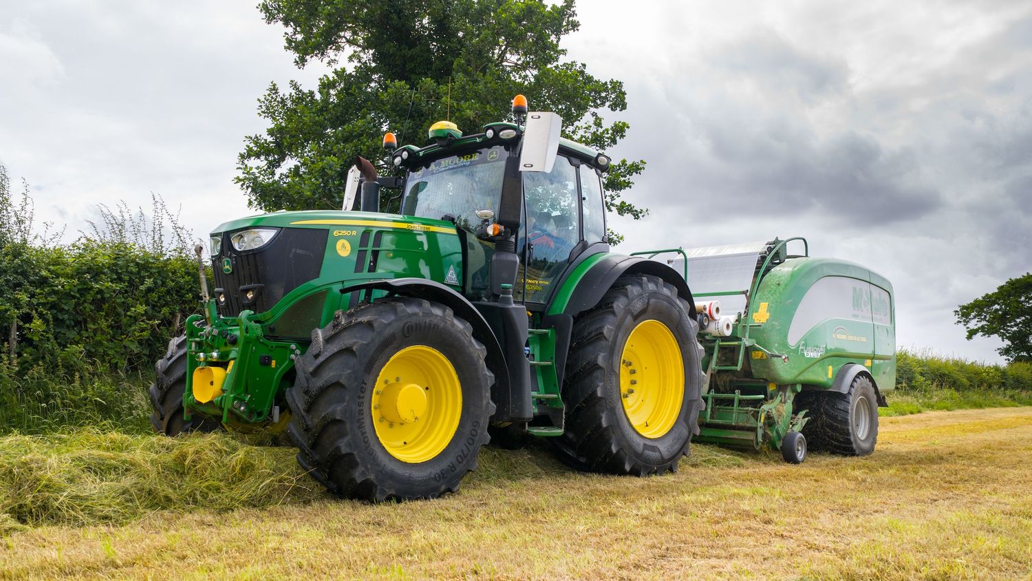 Modern green John Deere tractor pulling baling equipment across a sunny hay field.