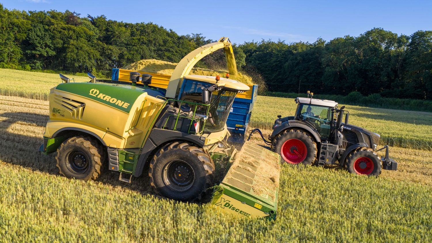 Agricultural machinery performs farming operations on a rural field under bright summer skies.