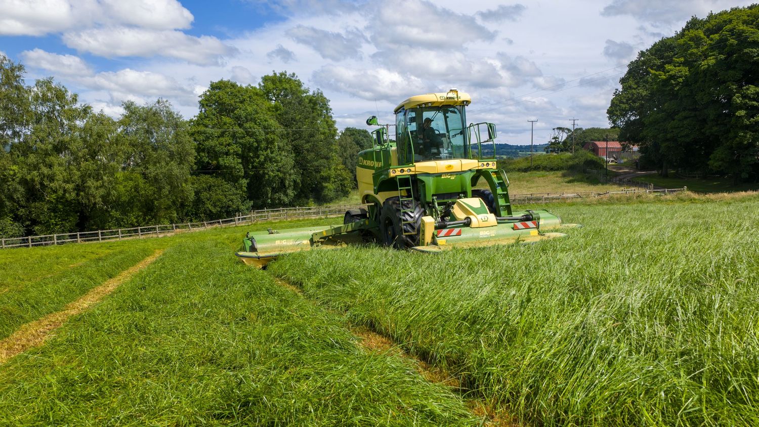 Large hay mowing tractor equipment operates in green farmland under blue cloudy skies creating rows of cut grass.