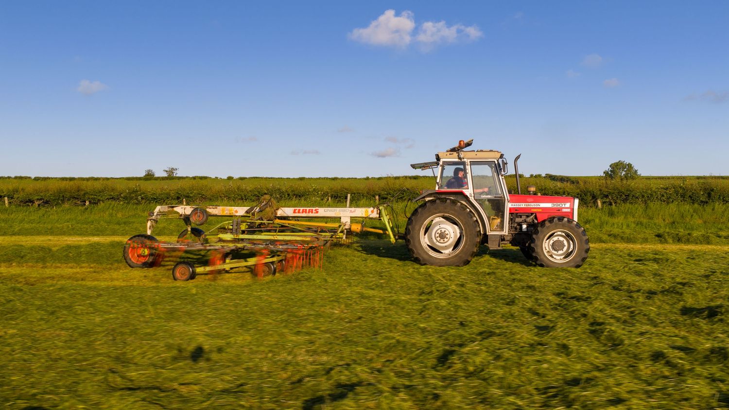 Red tractor with hay equipment working in a green agricultural field under blue summer sky.