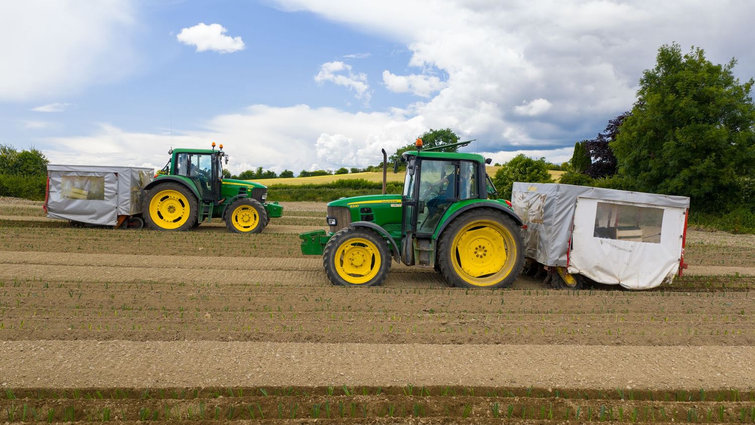 A panoramic view of farm fields with green John Deere tractors and agricultural equipment under a cloudy blue sky.
